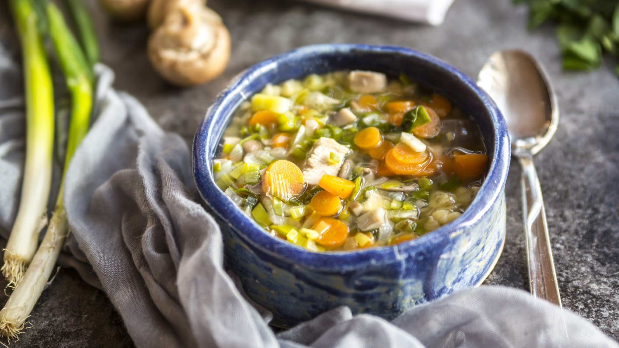 Draufsicht schräg: Ein Topf herbstliche Hühnersuppe auf einem Holztisch. Drum herum liegen frische Zutaten, ein Handtuch und ein Suppenlöffel.