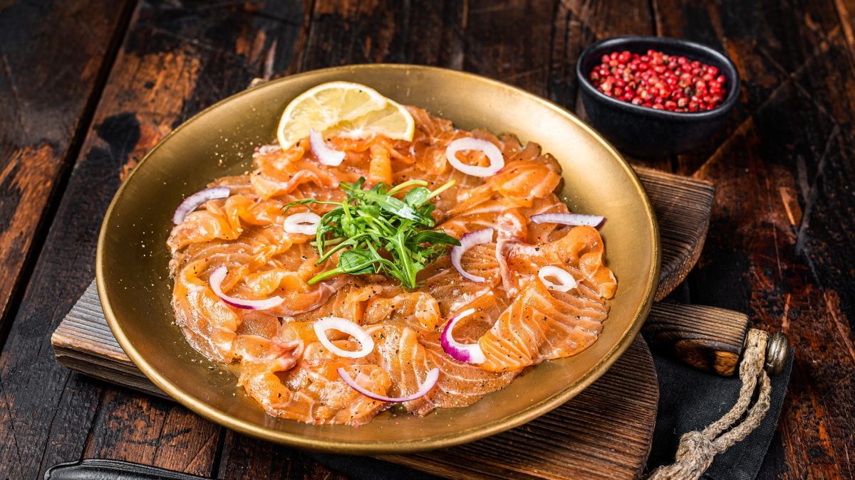 Draufsicht schräg: Ein goldfarbener Teller mit einem Lachs-Carpaccio mit Zitronen-Vinaigrette liegt auf einem Holzbrett, dass auf einem Holztisch liegt. Davor ist eine Gabel zu sehen. Im Hintergrund steht eine schwarze Schüssel mit rotem Pfeffer.