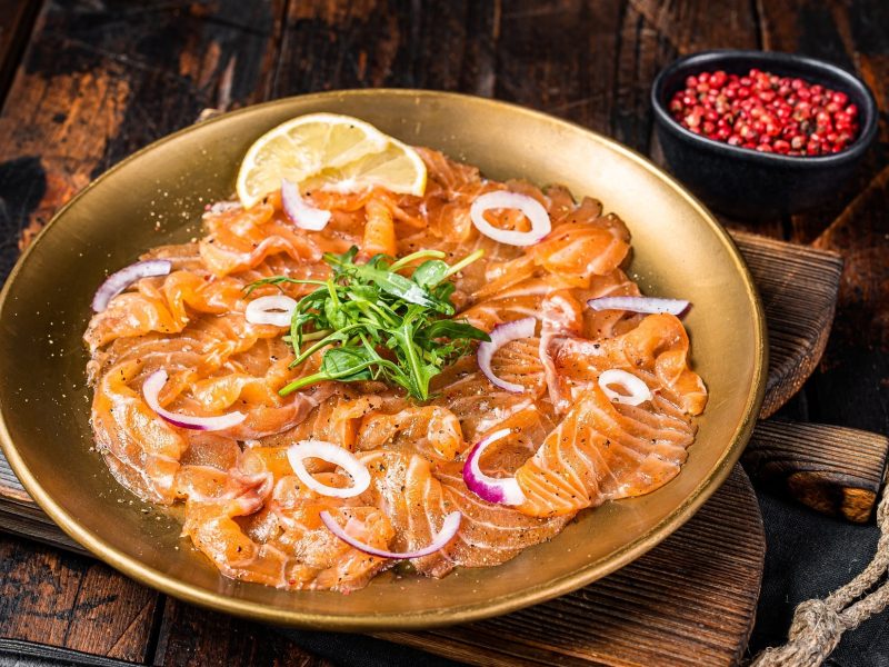 Draufsicht schräg: Ein goldfarbener Teller mit einem Lachs-Carpaccio mit Zitronen-Vinaigrette liegt auf einem Holzbrett, dass auf einem Holztisch liegt. Davor ist eine Gabel zu sehen. Im Hintergrund steht eine schwarze Schüssel mit rotem Pfeffer.