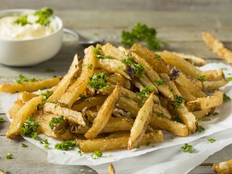 Leichte schräge Draufsicht: knusprige Parmesan-Pommes aus der Heißluftfritteuse liegen auf weißem Papier auf einem Untergrund aus Holz. Sie sind mit Petersilie bestreut. Im Hintergrund ist eine kleine weiße Schale mit Mayonnaise.