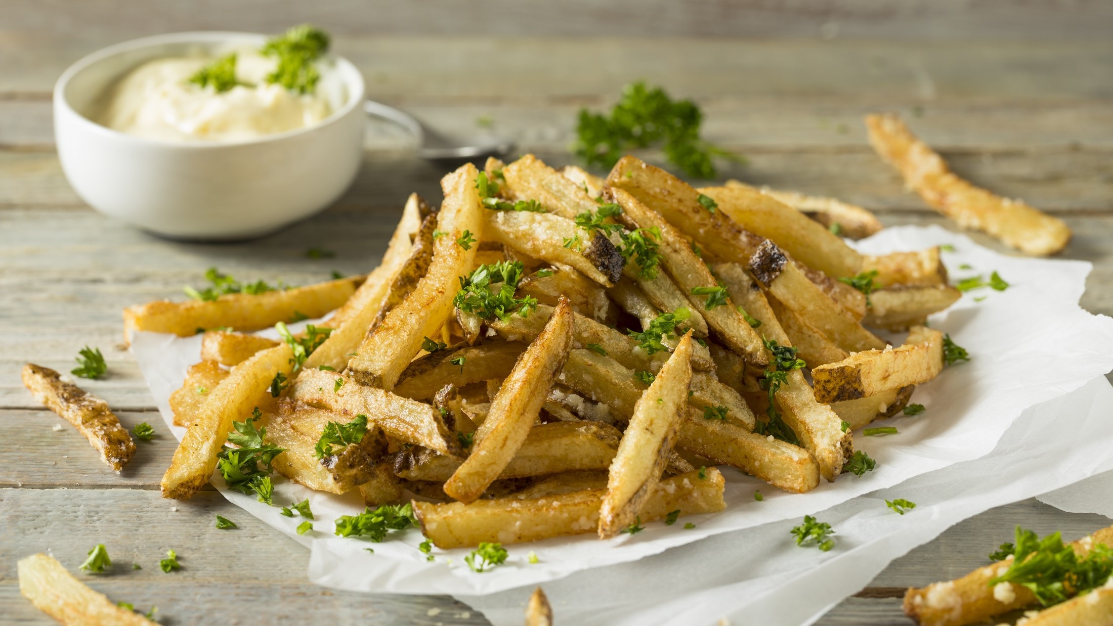 Leichte schräge Draufsicht: knusprige Parmesan-Pommes aus der Heißluftfritteuse liegen auf weißem Papier auf einem Untergrund aus Holz. Sie sind mit Petersilie bestreut. Im Hintergrund ist eine kleine weiße Schale mit Mayonnaise.