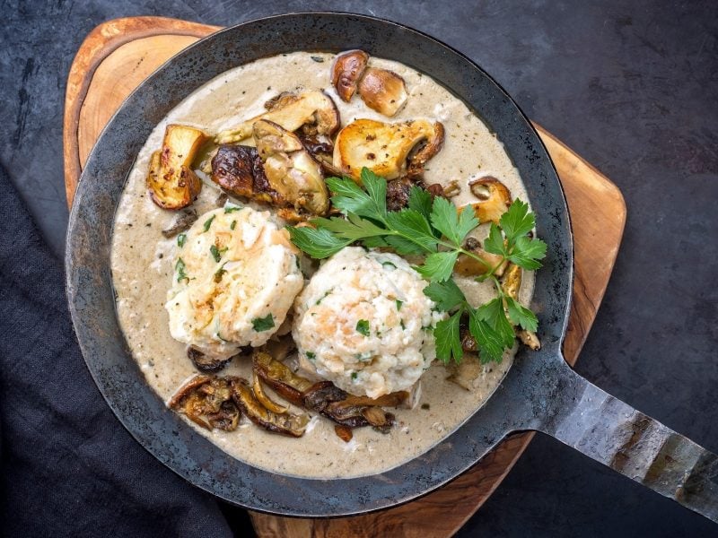 Draufsicht: Eine gußeiserne Pfanne Rahmschwammerl mit Knödel auf einem schwarzen Untergrund.