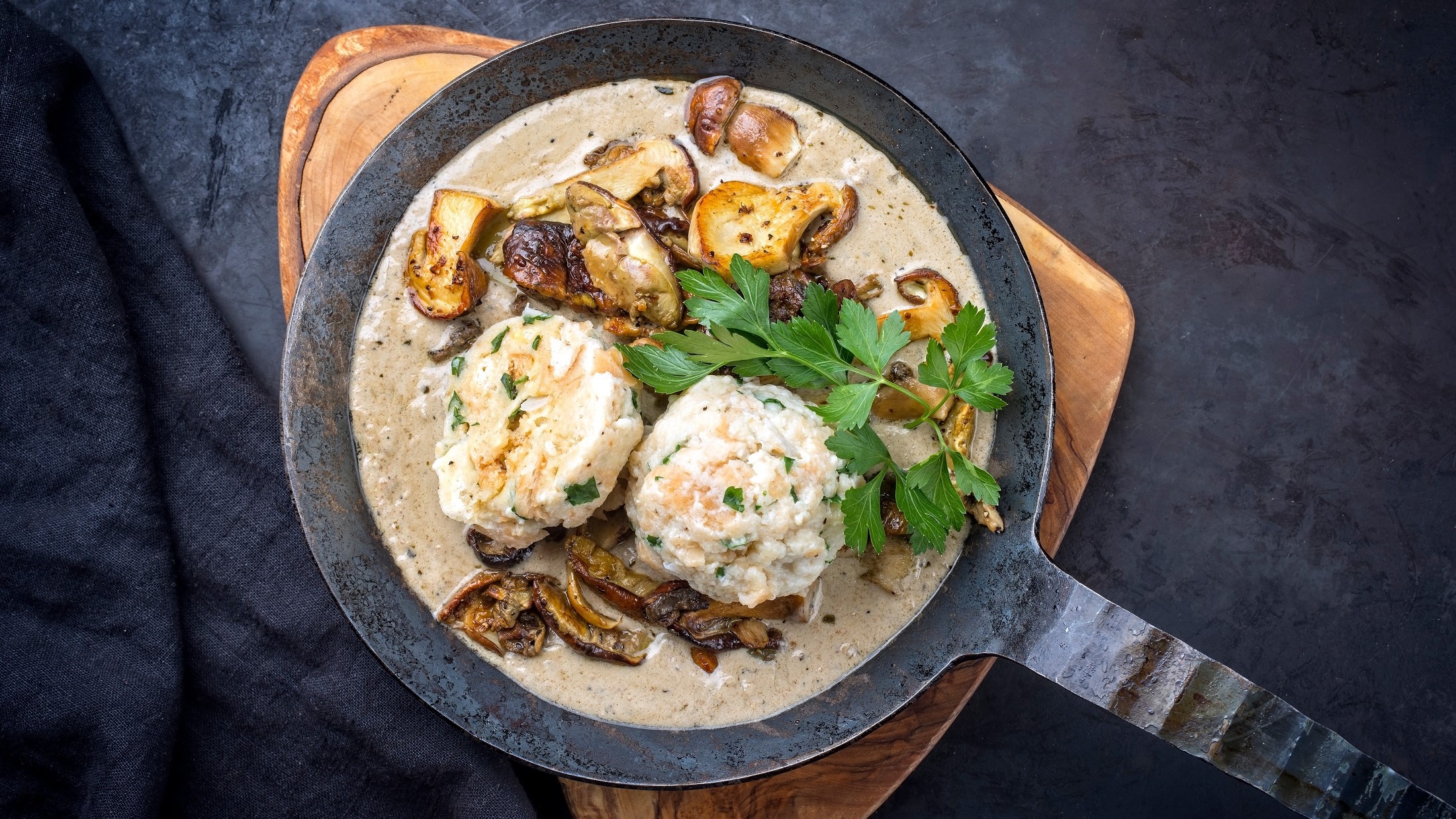 Draufsicht: Eine gußeiserne Pfanne Rahmschwammerl mit Knödel auf einem schwarzen Untergrund.