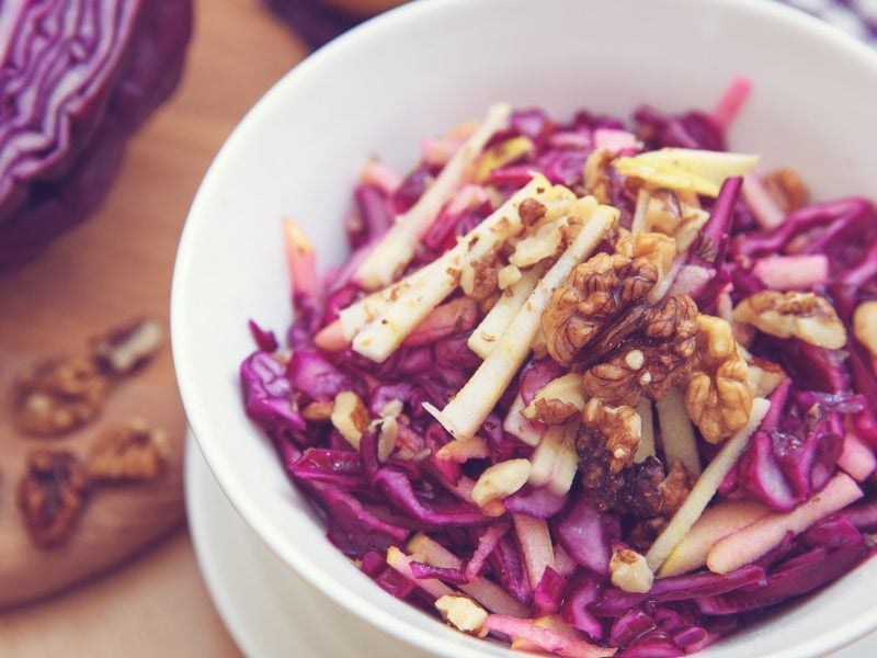 Draufsicht: Eine weiße Salatschale mit einem Rotkohlsalat mit Walnüssen und Äpfeln. Der steht auf einem Untergrund aus Holz. Drum herum liegen ein Tuch und ein angeschnittener Rotkohl.