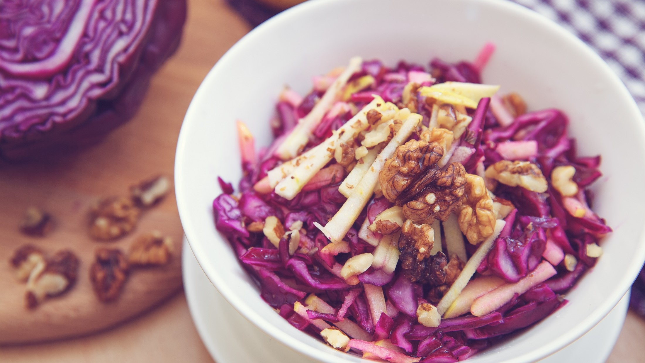 Draufsicht: Eine weiße Salatschale mit einem Rotkohlsalat mit Walnüssen und Äpfeln. Der steht auf einem Untergrund aus Holz. Drum herum liegen ein Tuch und ein angeschnittener Rotkohl.