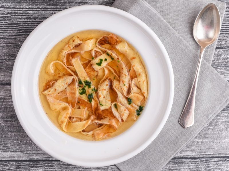 Flädlesuppe mit dünnen Pfannkuchenstreifen in einer klaren Brühe, serviert in einem weißen Teller auf grauer Holzoberfläche.