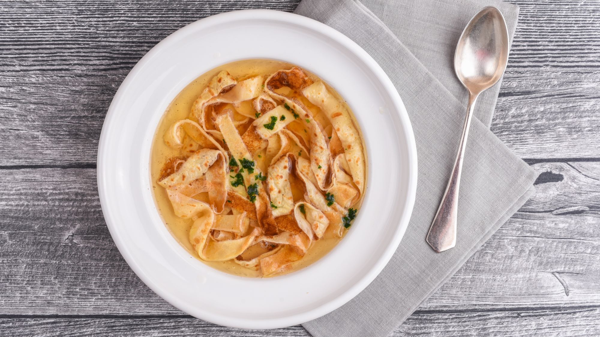 Flädlesuppe mit dünnen Pfannkuchenstreifen in einer klaren Brühe, serviert in einem weißen Teller auf grauer Holzoberfläche.