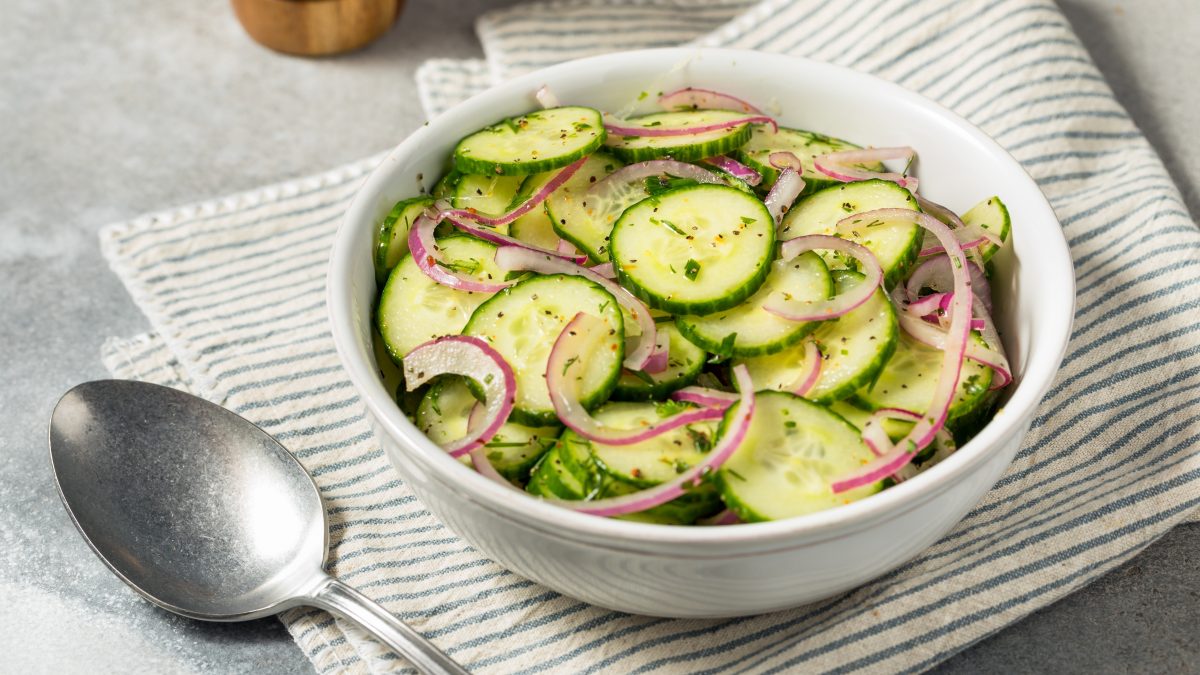 Gurkensalat mit roten Zwiebeln und Kräutern in einer weißen Schale auf gestreiftem Stofftuch, daneben ein silberner Löffel.