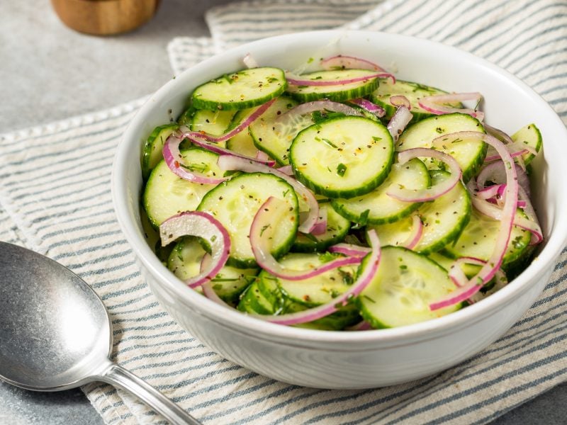 Gurkensalat mit roten Zwiebeln und Kräutern in einer weißen Schale auf gestreiftem Stofftuch, daneben ein silberner Löffel.