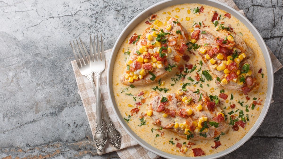 Drei Hähnchenbrustfilets in cremiger Sauce mit Mais, Speck und frischem Koriander auf einem grauen Marmortisch, daneben zwei silberne Gabeln.