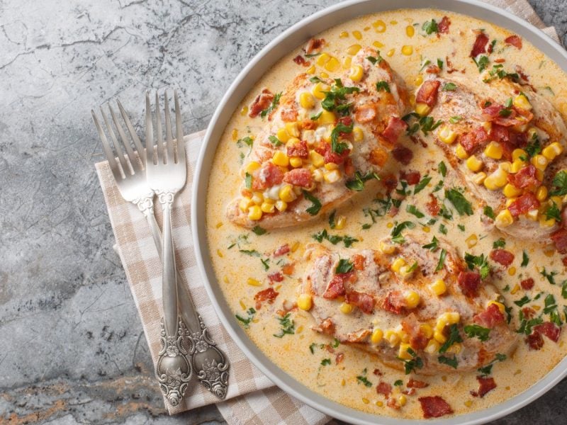 Drei Hähnchenbrustfilets in cremiger Sauce mit Mais, Speck und frischem Koriander auf einem grauen Marmortisch, daneben zwei silberne Gabeln.