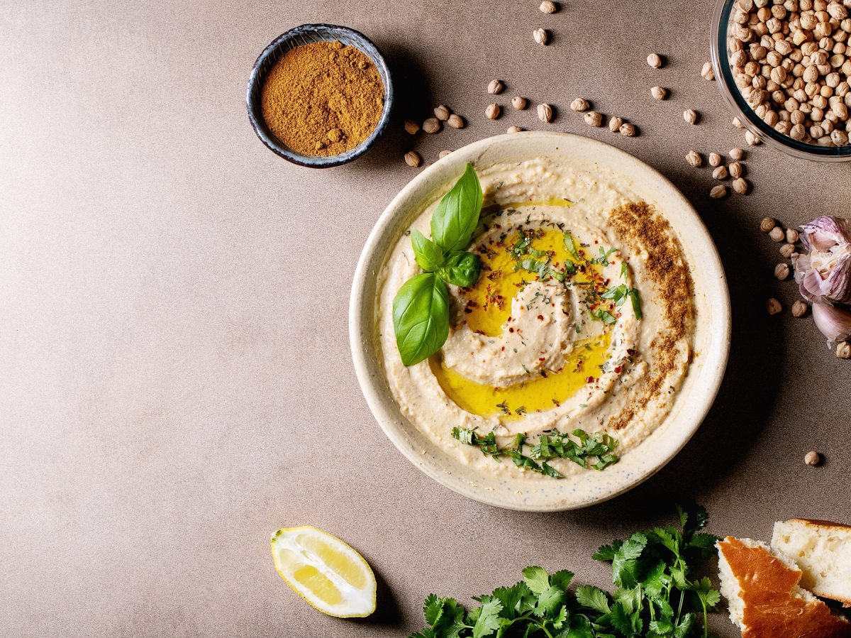 Eine Schale mit Hummus mit Olivenöl und Basilikum, daneben Zutaten für Hummus von oben fotografiert.