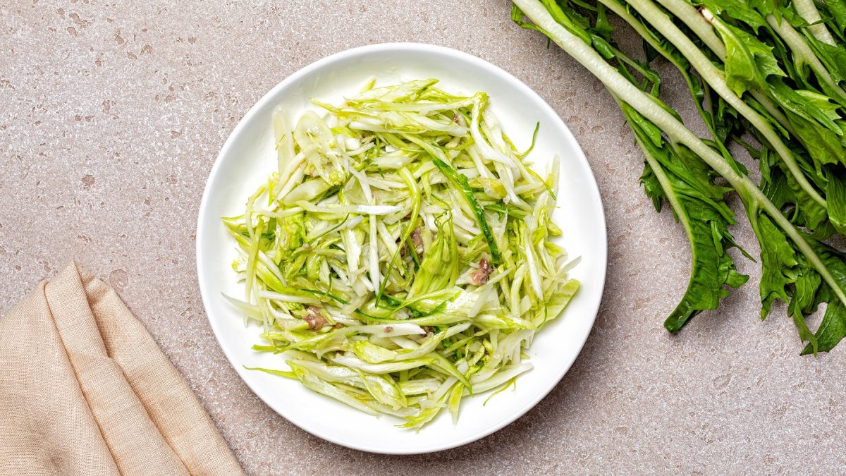 Insalata di puntarelle auf einem weißen Teller, daneben ein frischer Bund Puntarelle. Von oben fotografiert.