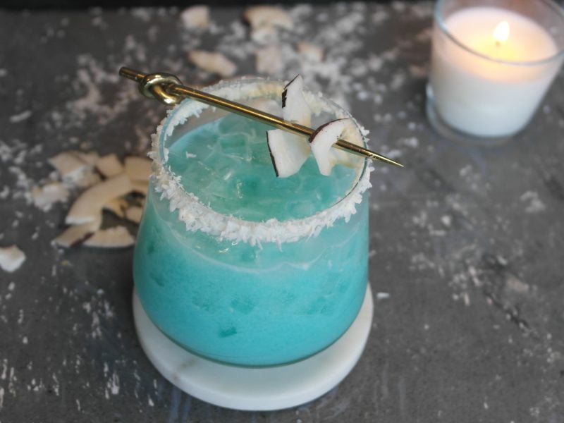 Der blaue Cocktail auf grauem Steintisch mit Kokosraspeln und Kokoschips sowie einer weißen Kerze im Hintergrund.