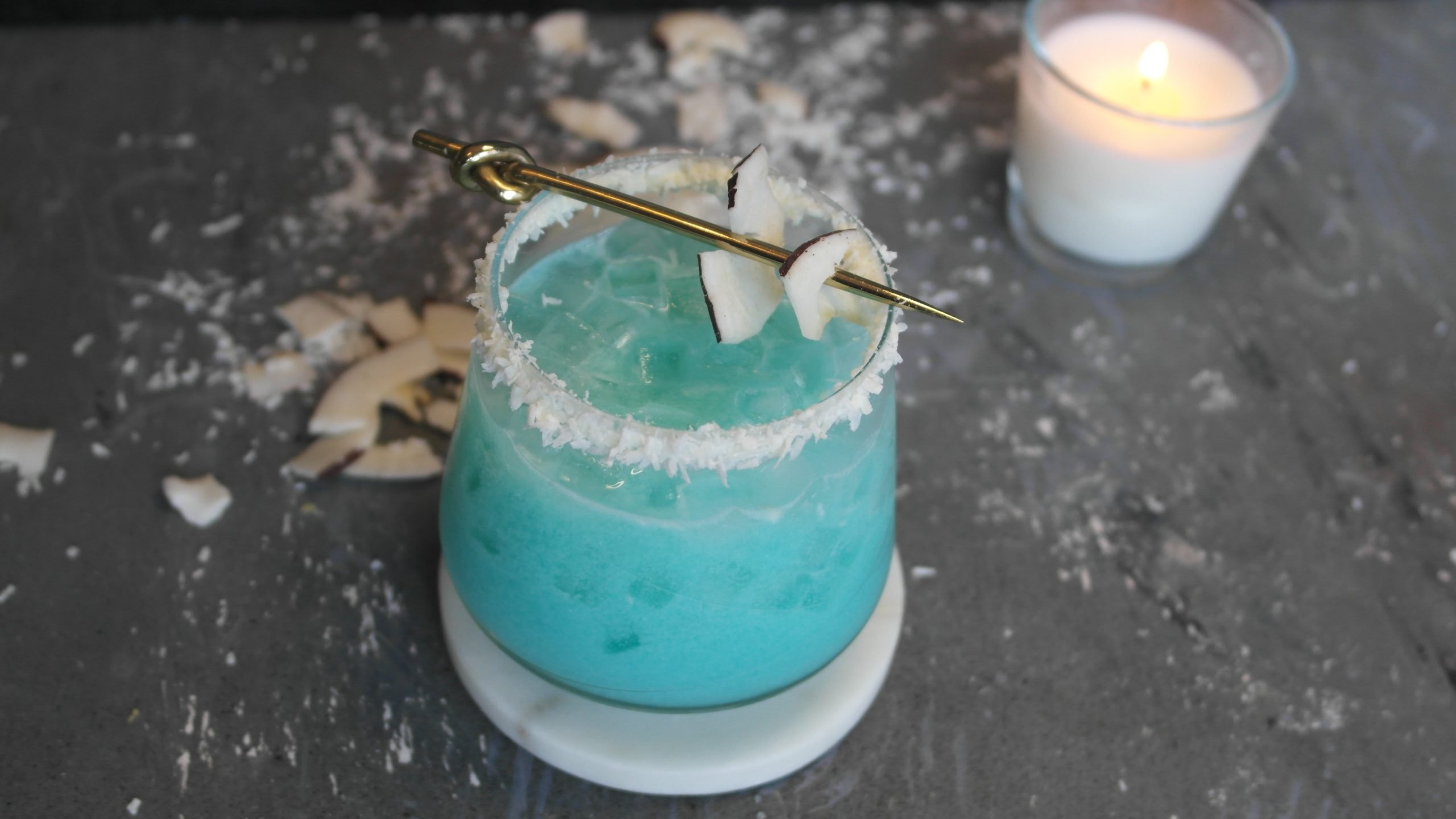 Der blaue Cocktail auf grauem Steintisch mit Kokosraspeln und Kokoschips sowie einer weißen Kerze im Hintergrund.