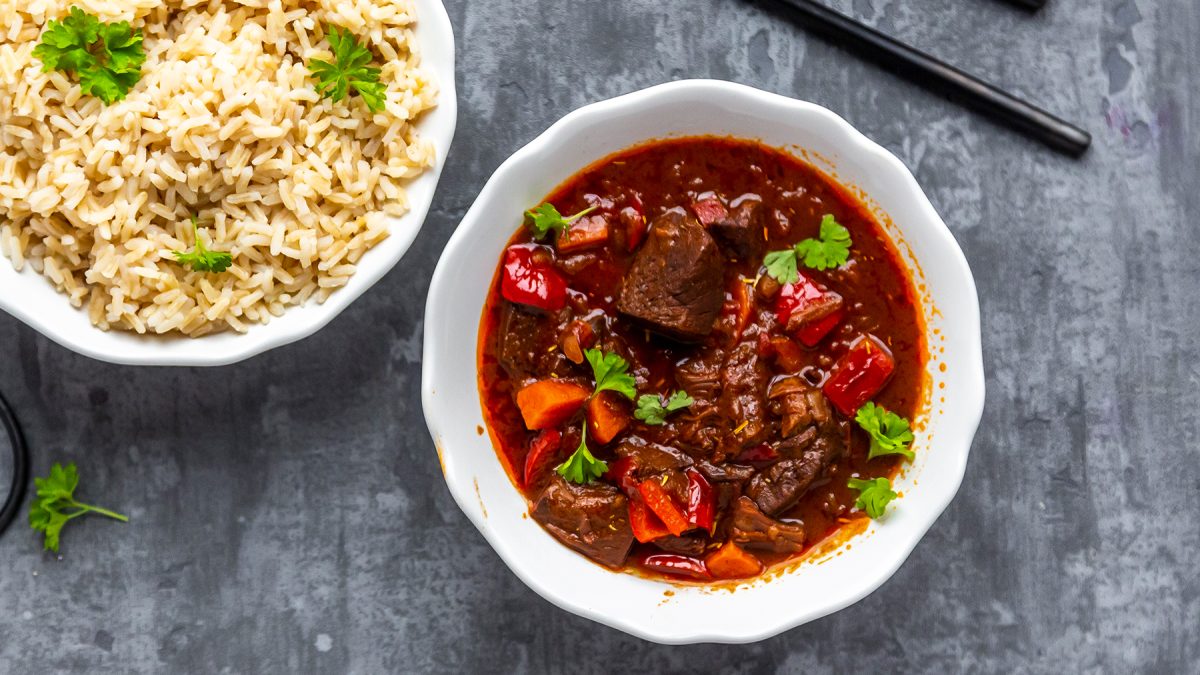 Veganes Gulasch mit Jackfruit in einer weißen Schüssel, links davon eine weiße Schüssel mit Reis. Der Hintergrund ist grau.