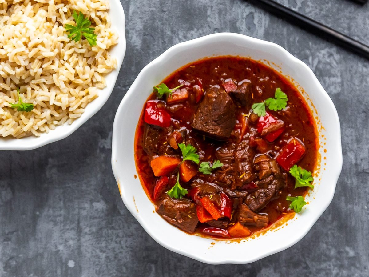 Veganes Gulasch mit Jackfruit in einer weißen Schüssel, links davon eine weiße Schüssel mit Reis. Der Hintergrund ist grau.