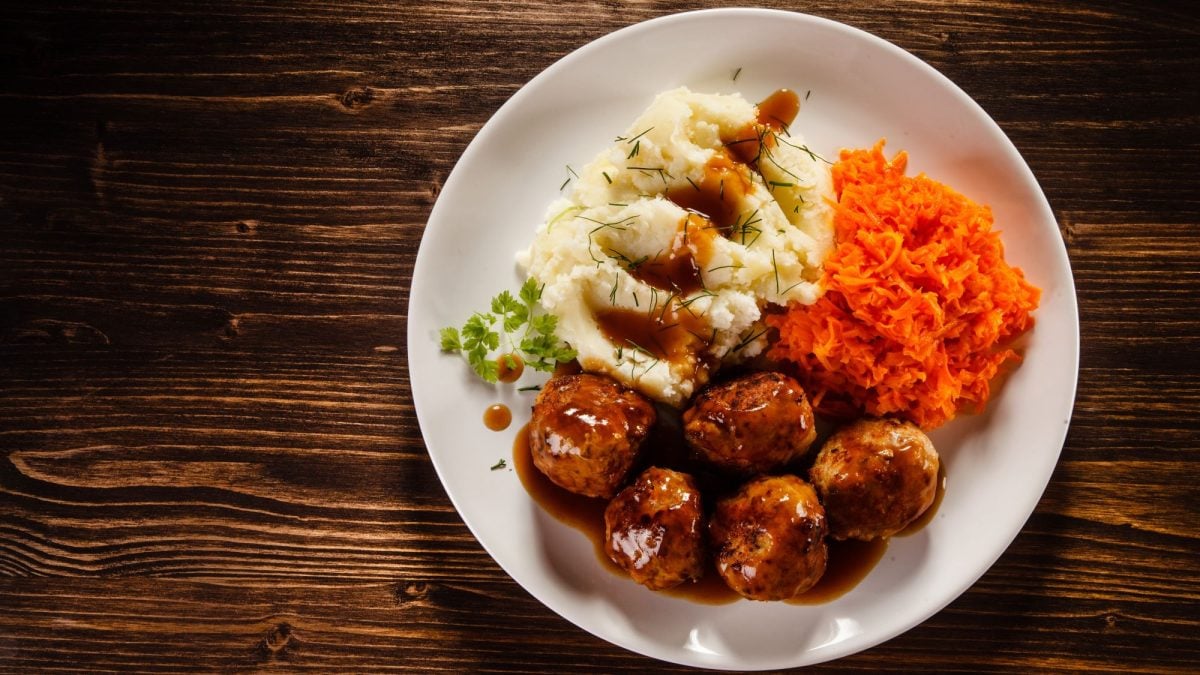 Teller mit polnischen Hackbällchen "Klopsy", Kartoffelpüree und geraspelten Möhren in Soße auf dunklem Holztisch, garniert mit Kräutern.