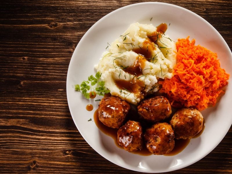 Teller mit polnischen Hackbällchen "Klopsy", Kartoffelpüree und geraspelten Möhren in Soße auf dunklem Holztisch, garniert mit Kräutern.