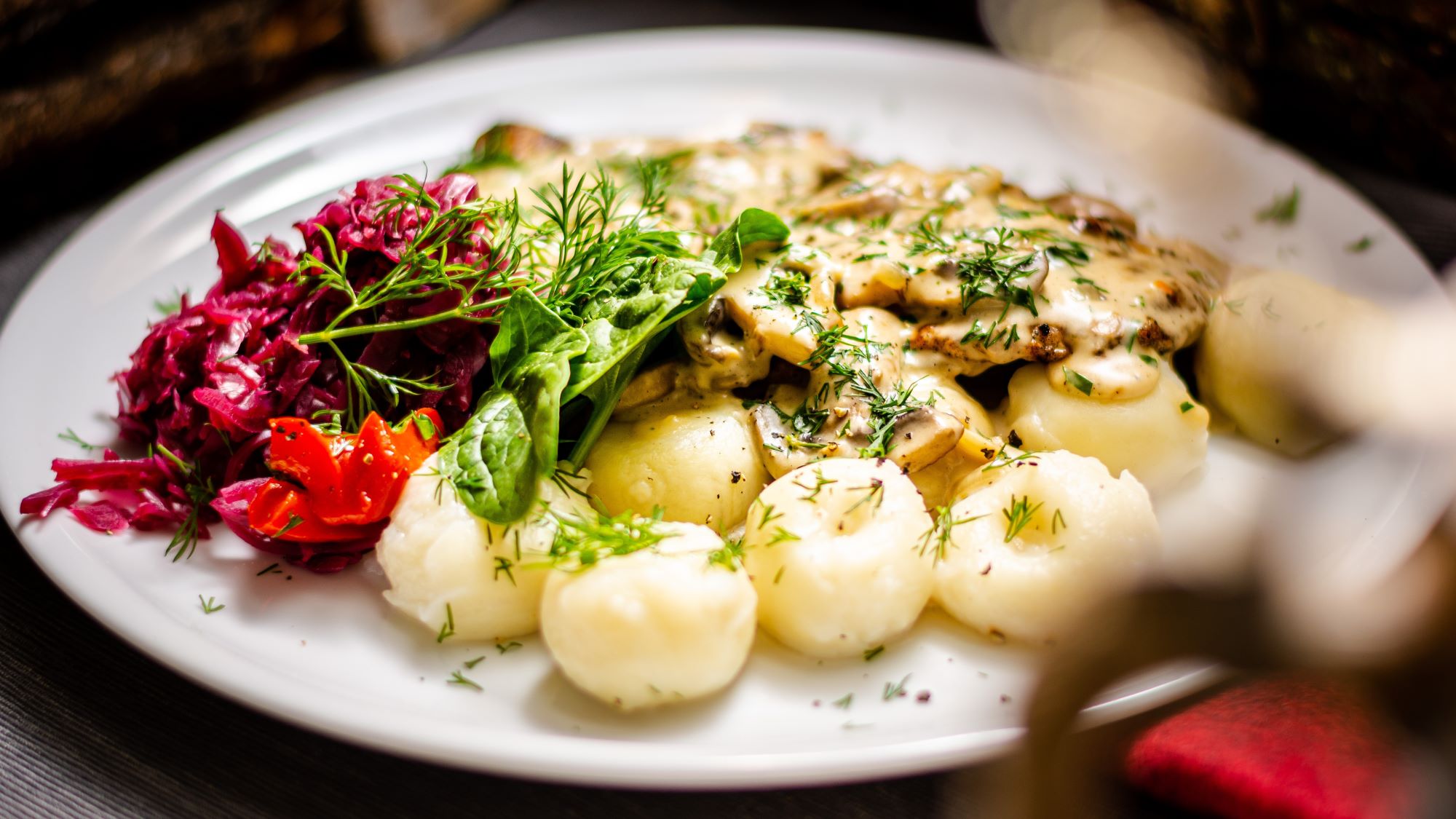 Teller mit schlesischen Kartoffelklößen "Kluski śląskie", überzogen mit Pilzsoße und frischem Dill, dazu Rote Bete Salat und Spinatblätter.