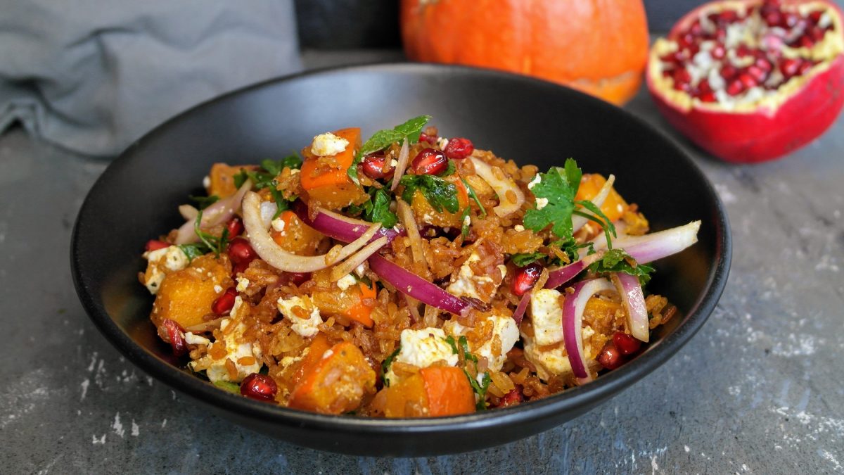 Eine schwarze Schale mit dem knusprigen Reissalat auf einem Steintisch mit Kürbis und Granatapfel im Hintergrund. Alles von der Seite fotografiert.