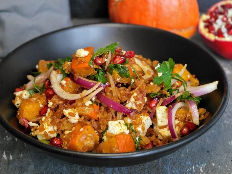 Eine schwarze Schale mit dem knusprigen Reissalat auf einem Steintisch mit Kürbis und Granatapfel im Hintergrund. Alles von der Seite fotografiert.