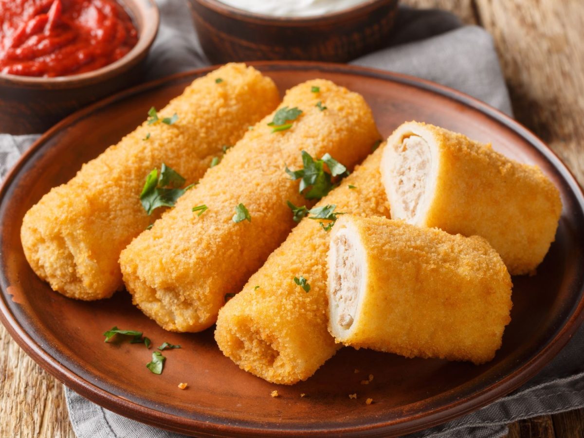 Vier knusprige gefüllte Pfannkuchen, auch polnische Kroketten genannt "Korkiety" auf braunem Teller, dazu Tomatensoße und Sauerrahm in rustikalen Schälchen.