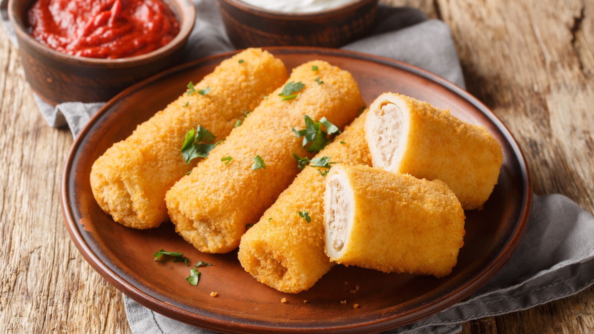 Vier knusprige gefüllte Pfannkuchen, auch polnische Kroketten genannt "Korkiety" auf braunem Teller, dazu Tomatensoße und Sauerrahm in rustikalen Schälchen.