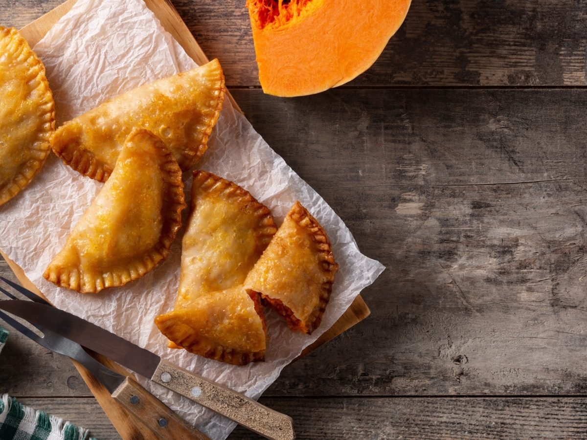 Fünf Kürbis-Empanadas von oben fotografiert auf einem Holzbrett mit Papier, daneben Küchentuch und angeschnittener Kürbis.