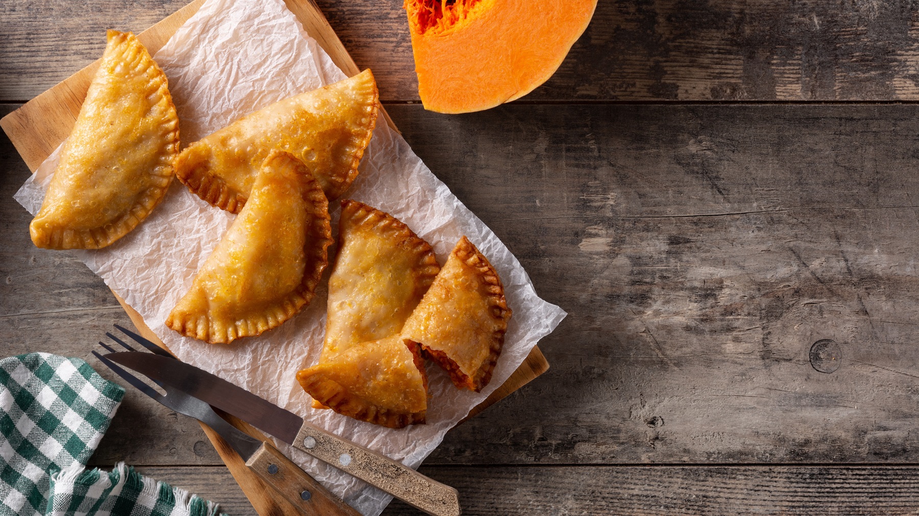 Fünf Kürbis-Empanadas von oben fotografiert auf einem Holzbrett mit Papier, daneben Küchentuch und angeschnittener Kürbis.