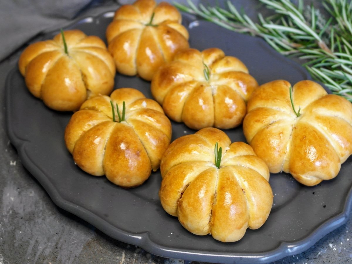 Sechs Kürbisbrötchen auf einem grauen Teller und Steintisch von schräg oben fotografiert. Dahinter frische Kräuter und ein graues Tuch.