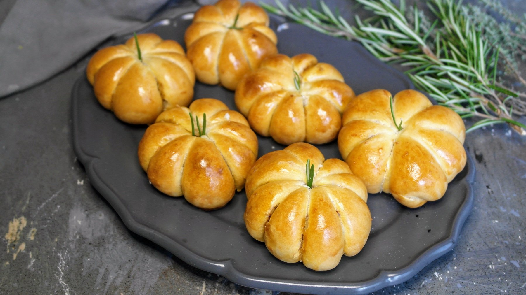 Sechs Kürbisbrötchen auf einem grauen Teller und Steintisch von schräg oben fotografiert. Dahinter frische Kräuter und ein graues Tuch.