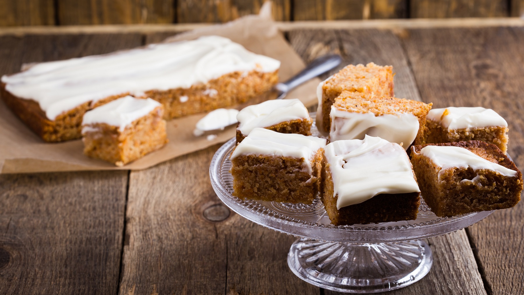 Eine Glasplatte mit einigen Stücken des Kuchens auf einem rustikalen Holztisch mit dem Blech und Kuchen im Hintergrund.