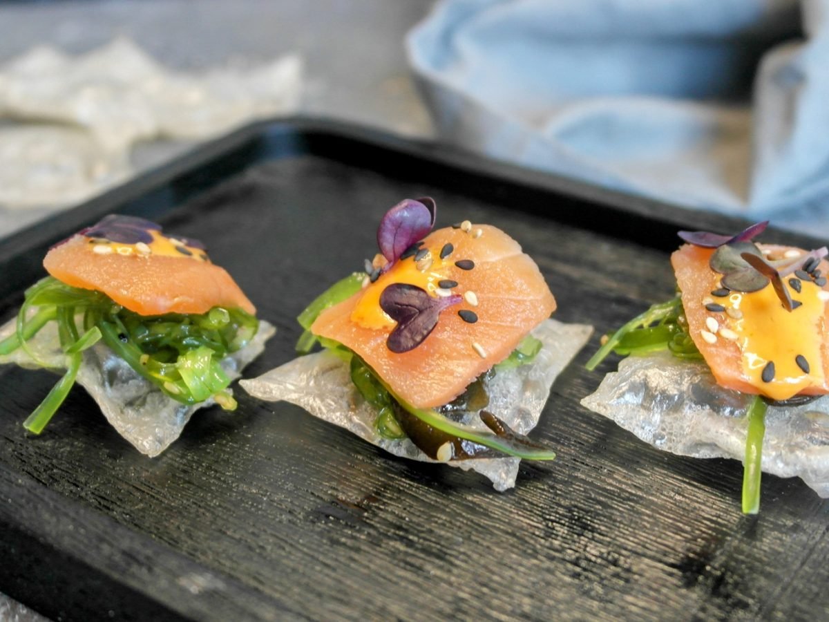 Ein schwarzes Brett mit drei Lachs-Häppchen auf einem grauen Steintisch von schräg oben fotografiert. Dahinter ein graues Tuch und Reispapierchips.