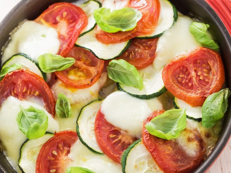 Frisches Tomaten-Zucchini-Gratin mit geschmolzenem Käse und Basilikumblättern in einer roten Auflaufform auf kariertem Tuch.