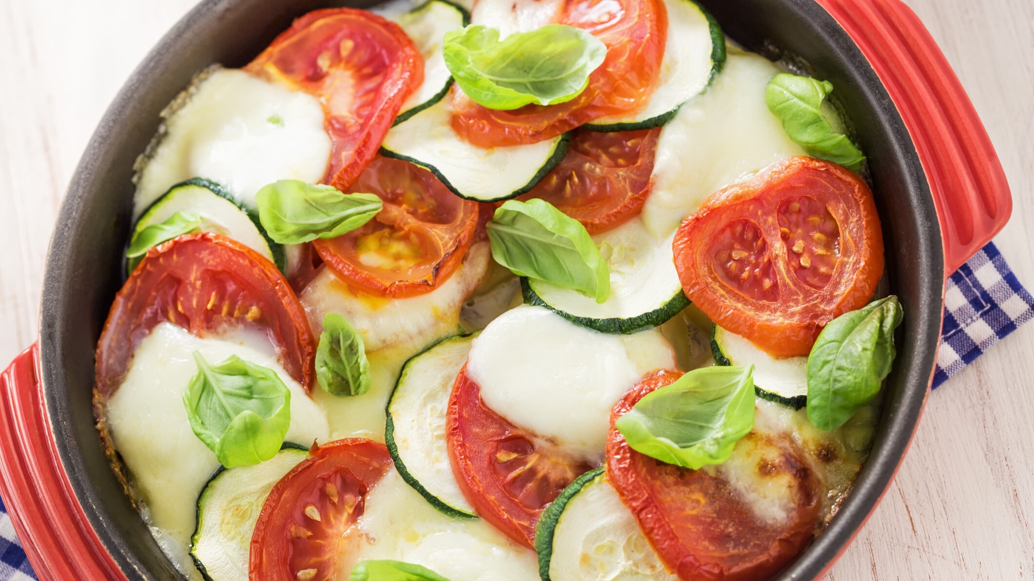 Frisches Tomaten-Zucchini-Gratin mit geschmolzenem Käse und Basilikumblättern in einer roten Auflaufform auf kariertem Tuch.