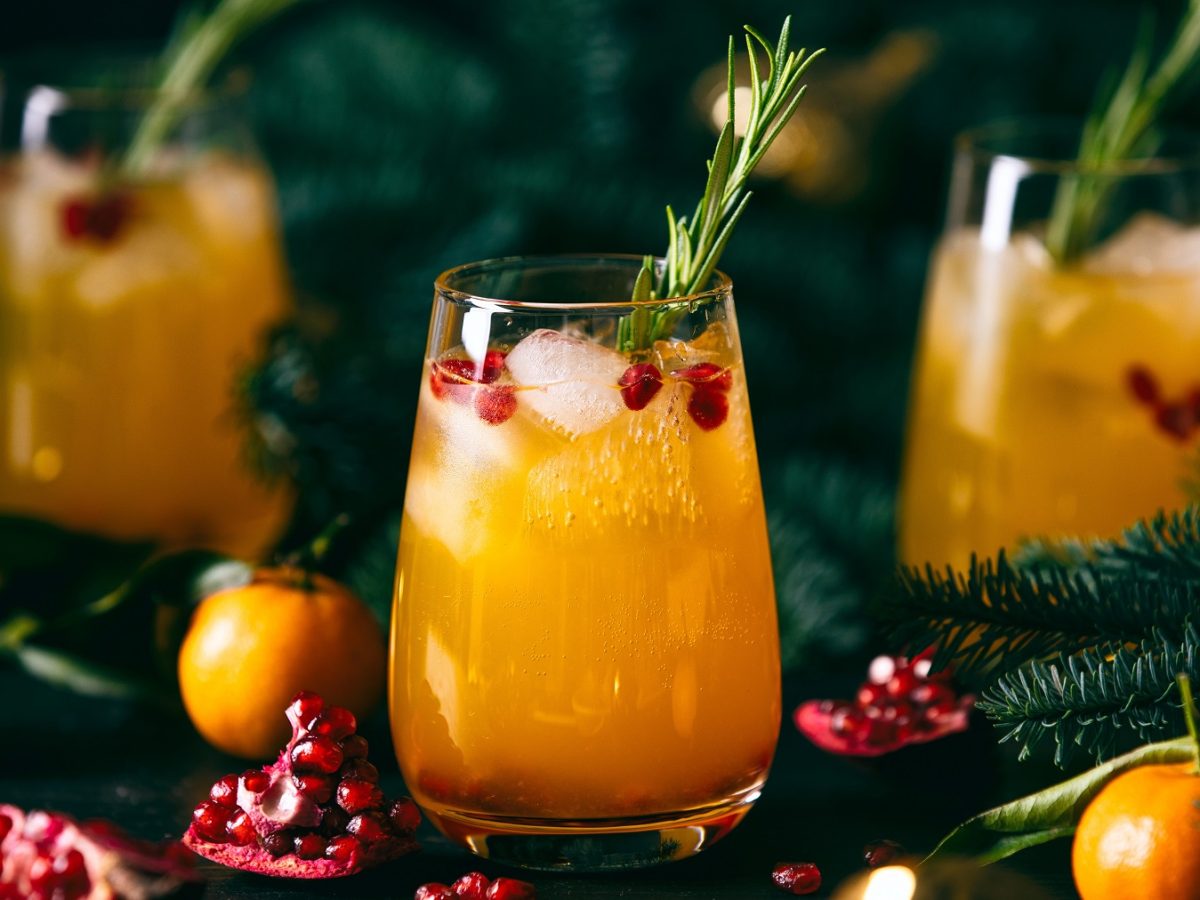 Drei Gläser mit Mandarinen-Granatapfel-Fizz umgeben von weihnachtlicher Deko sowie Granatäpfeln und Mandarinen.