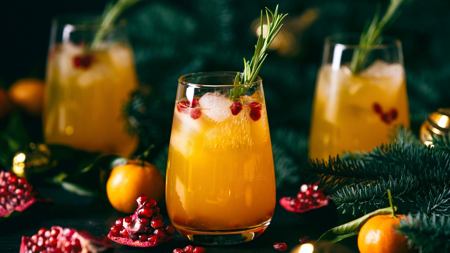 Drei Gläser mit Mandarinen-Granatapfel-Fizz umgeben von weihnachtlicher Deko sowie Granatäpfeln und Mandarinen.