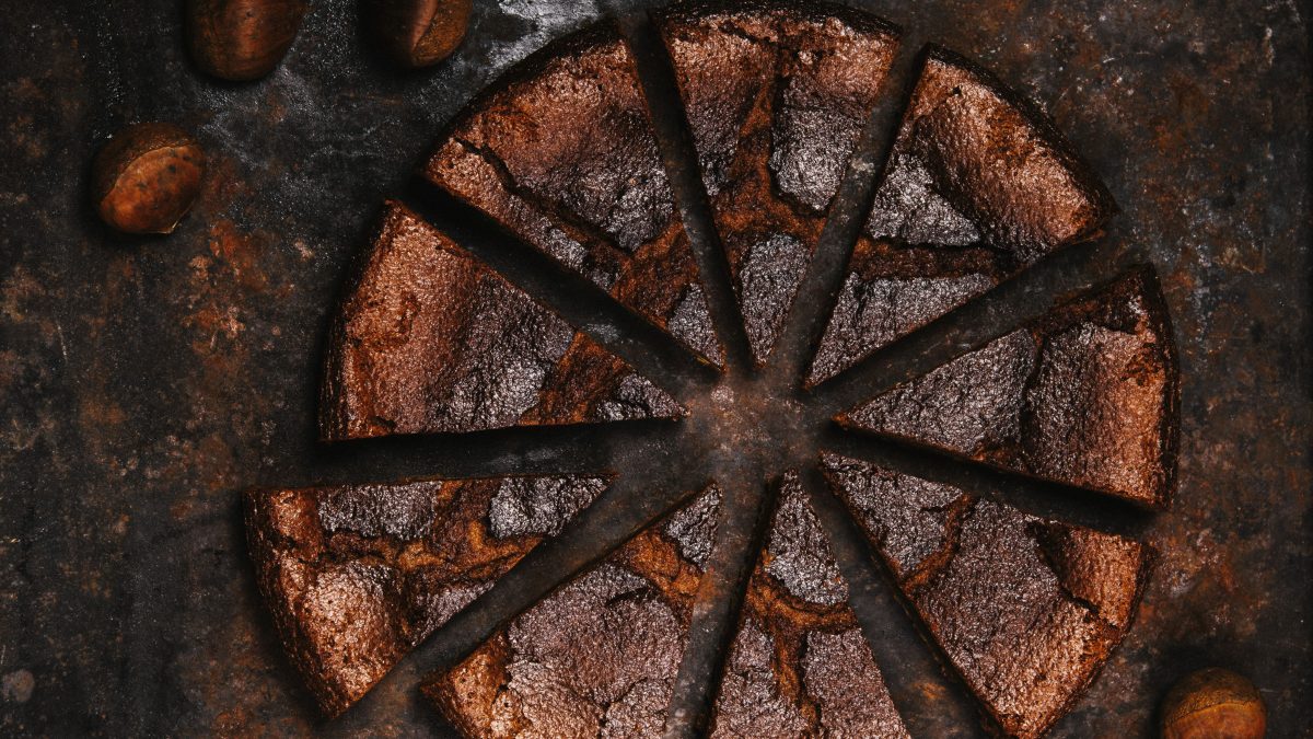 Der dunkelbraune Maronenkuchen von oben in kleinen Stücken geschnitten auf einem braunen Steinuntergrund fotografiert. Drumherum einige Maronen verteilt als Dekoration.