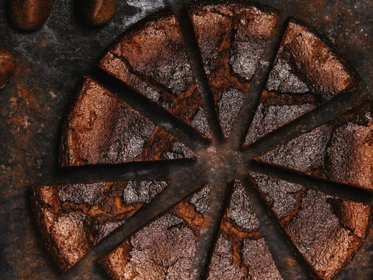 Der dunkelbraune Maronenkuchen von oben in kleinen Stücken geschnitten auf einem braunen Steinuntergrund fotografiert. Drumherum einige Maronen verteilt als Dekoration.