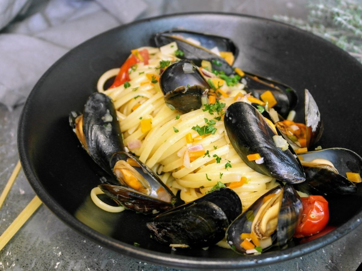 Ein schwarzer, tiefer Teller mit der Pasta auf einem grauen Steintisch mit ungekochter Pasta und einem grauen Tuch. Alles von schräg oben fotografiert.