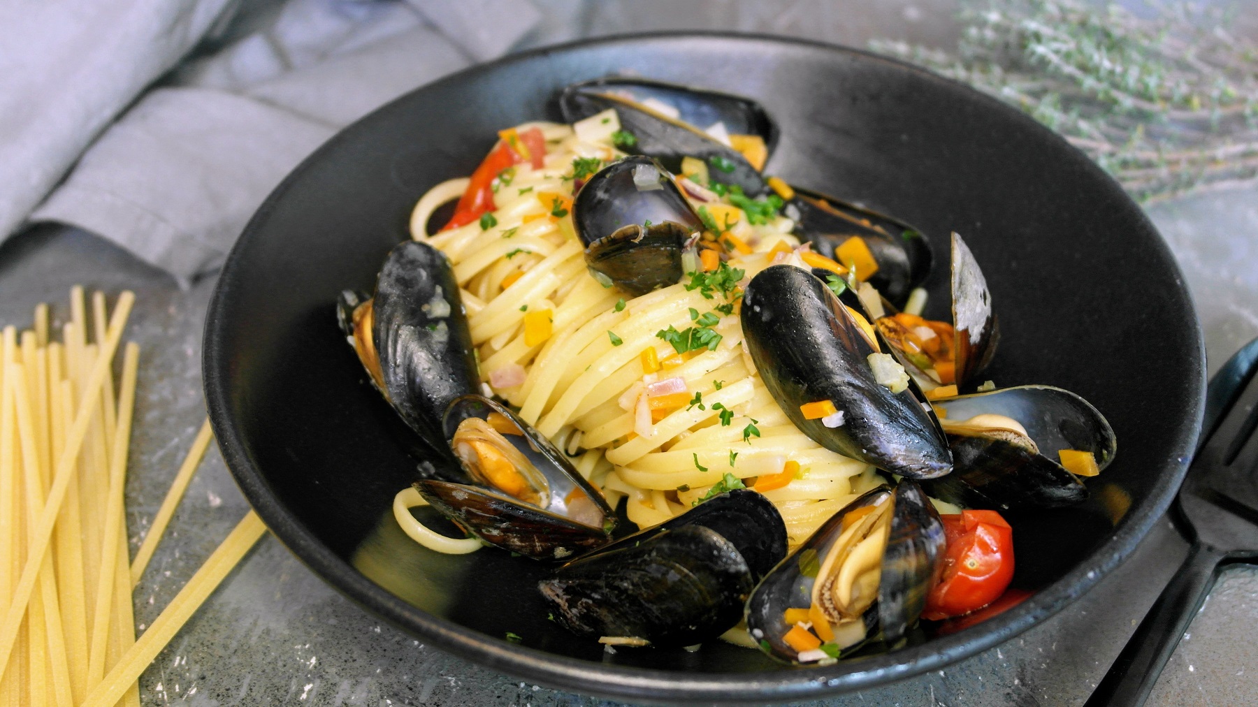 Ein schwarzer, tiefer Teller mit der Pasta auf einem grauen Steintisch mit ungekochter Pasta und einem grauen Tuch. Alles von schräg oben fotografiert.