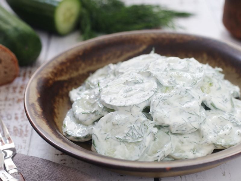 Polnischer Gurkensalat "Mizeria" mit frischem Dill in einer rustikalen Schale, daneben Gurken und Brot auf hellem Holztisch.