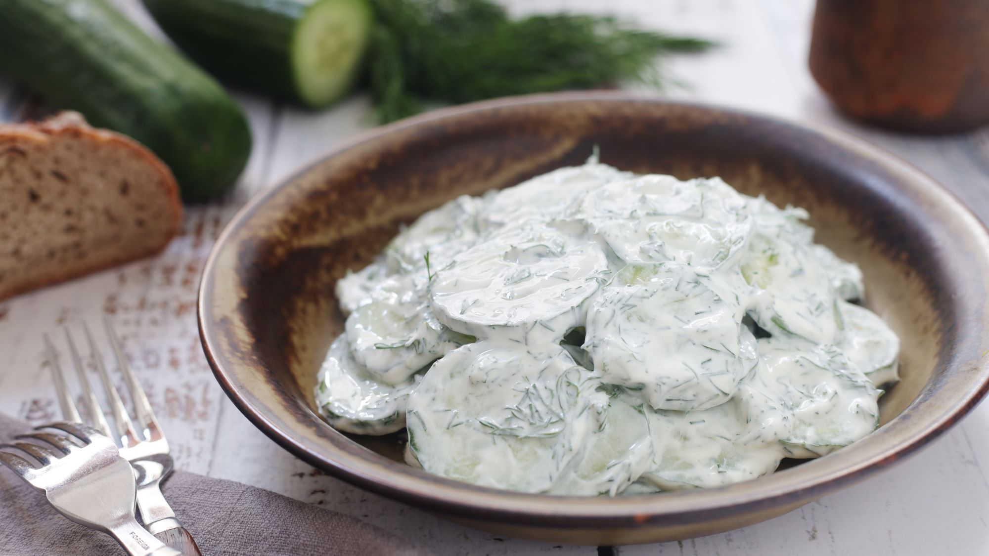 Polnischer Gurkensalat "Mizeria" mit frischem Dill in einer rustikalen Schale, daneben Gurken und Brot auf hellem Holztisch.