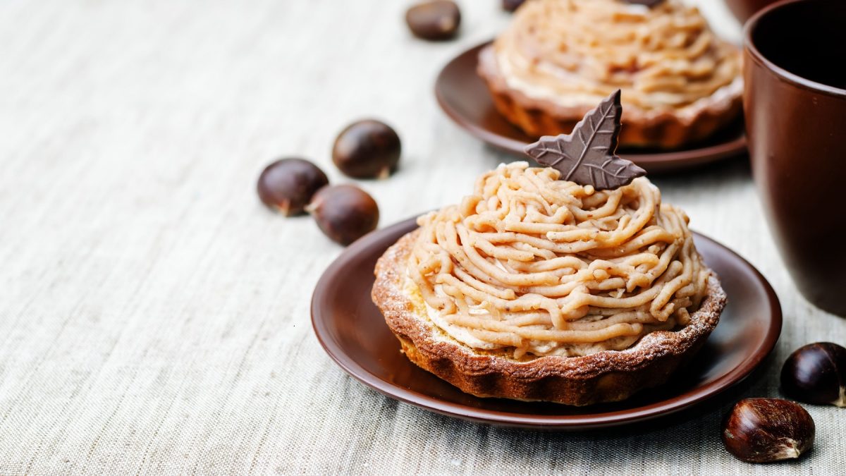 Zwei Mont Blanc Tartelettes von vorne forografiert, dekoriert mit einem Schokotäfelchen in Blattform. Daneben frische Maronen.