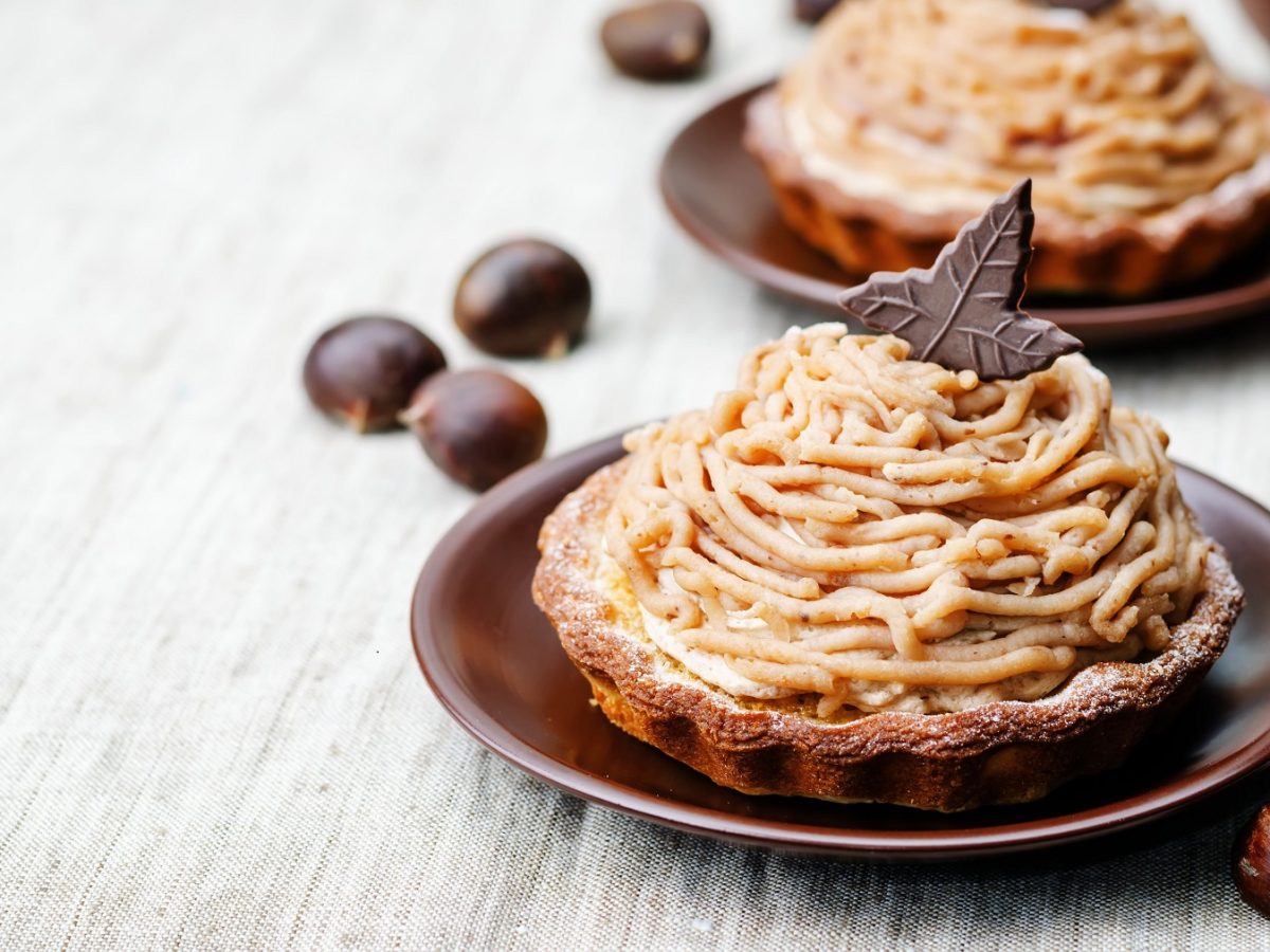 Zwei Mont Blanc Tartelettes von vorne forografiert, dekoriert mit einem Schokotäfelchen in Blattform. Daneben frische Maronen.