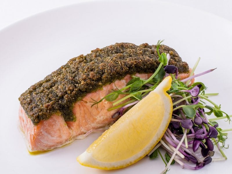 Ein weißer Teller mit dem Ofenlachs, einer Zitronenecke und Kresse auf weißem Unter- und Hintergrund fotografiert.