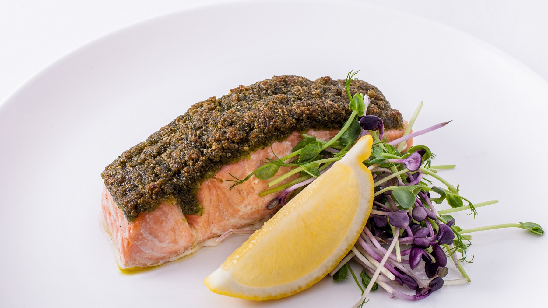 Ein weißer Teller mit dem Ofenlachs, einer Zitronenecke und Kresse auf weißem Unter- und Hintergrund fotografiert.