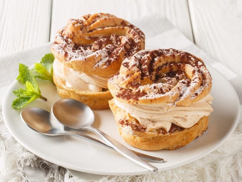 Zwei Paris-Brest mit Kakao-Topping auf einem weißen Teller schräg von oben fotografiert.