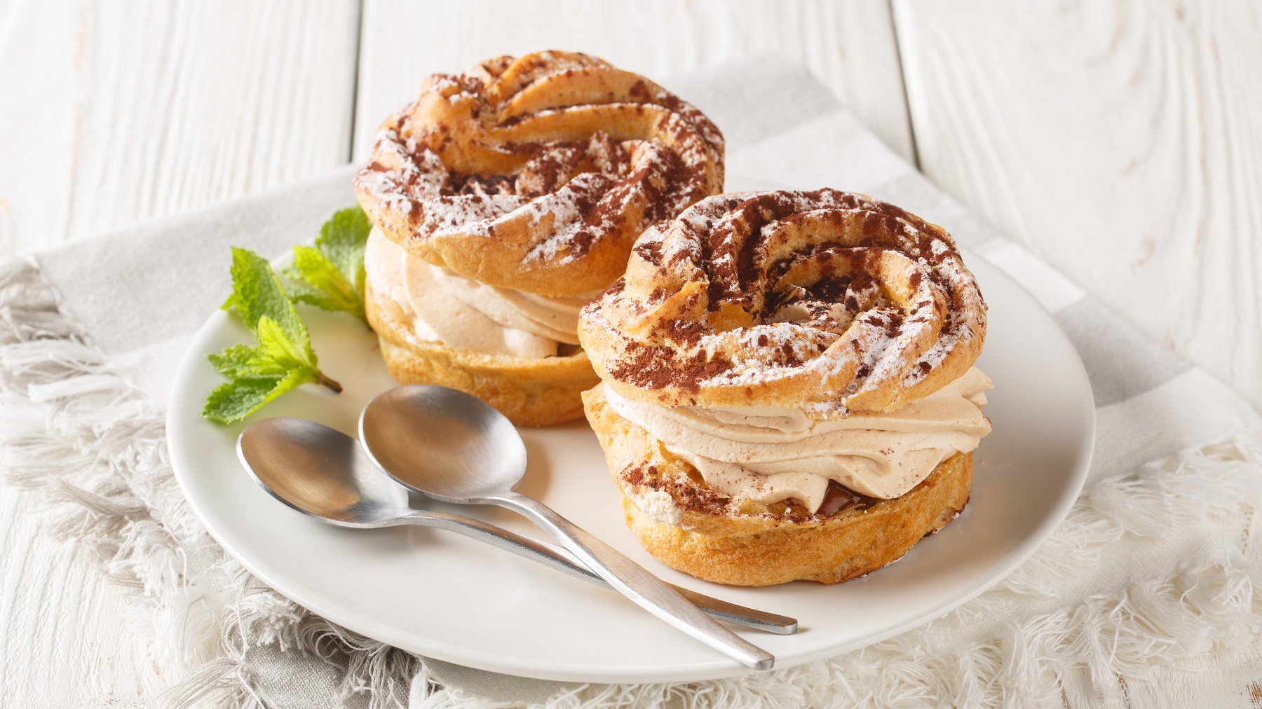 Zwei Paris-Brest mit Kakao-Topping auf einem weißen Teller schräg von oben fotografiert.